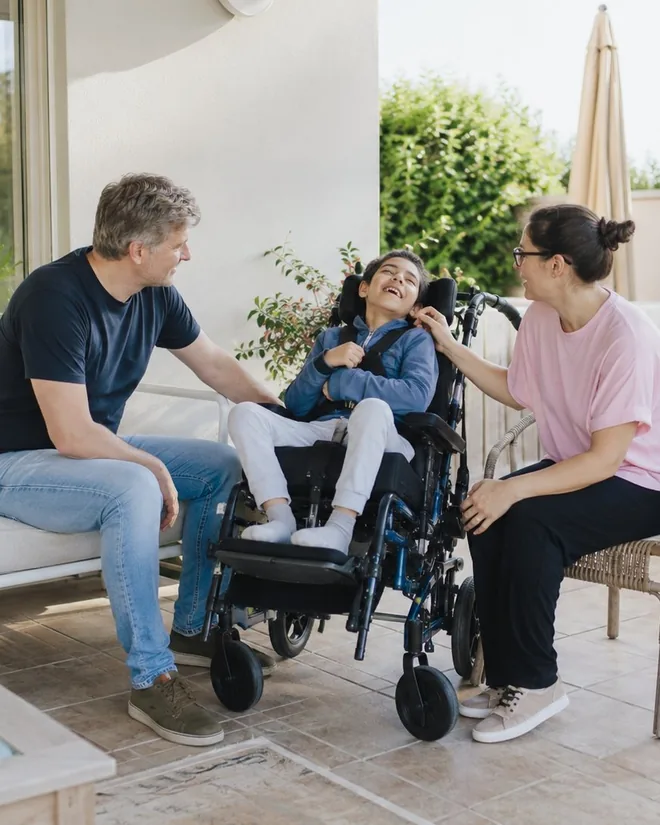 Accompagnement du handicap à domicile : un enfant en fauteuil roulant souriant, entouré de ses deux parents sur une terrasse