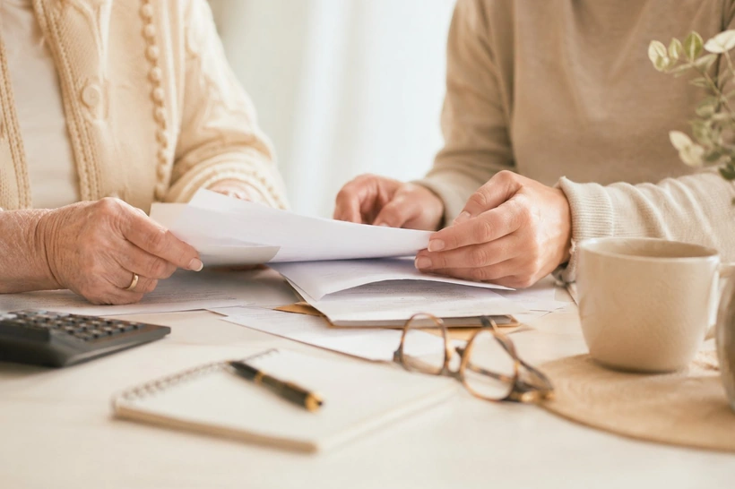Mains d'une personne âgée et d'un proche comparant des papiers, une calculatrice posée sur la table