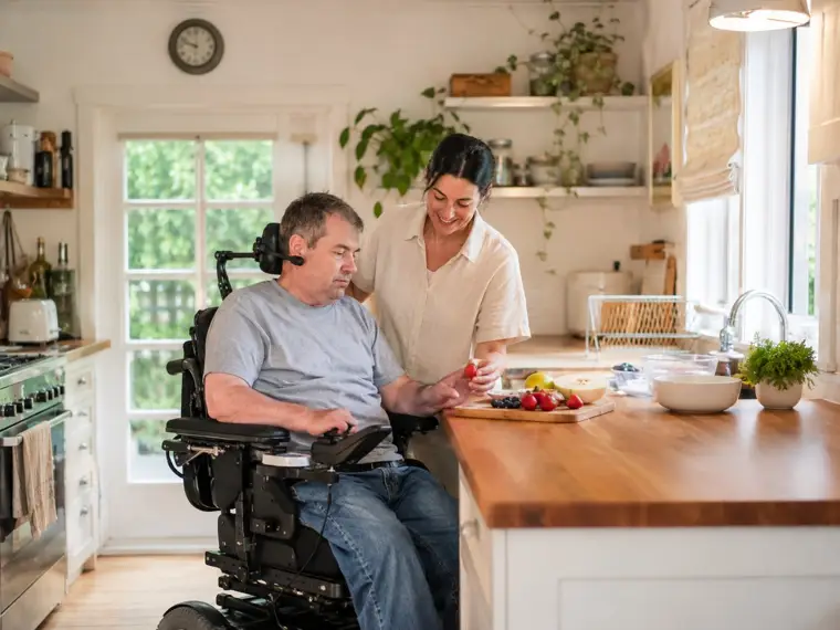 Un homme en fauteuil roulant électrique partage un moment dans sa cuisine avec son auxiliaire de vie AUXI'life